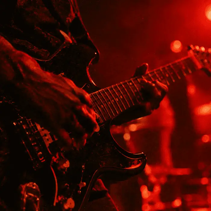 Close das mãos de um guitarrista da Maré Vermelha tocando em um show, com luzes de palco desfocadas ao fundo
